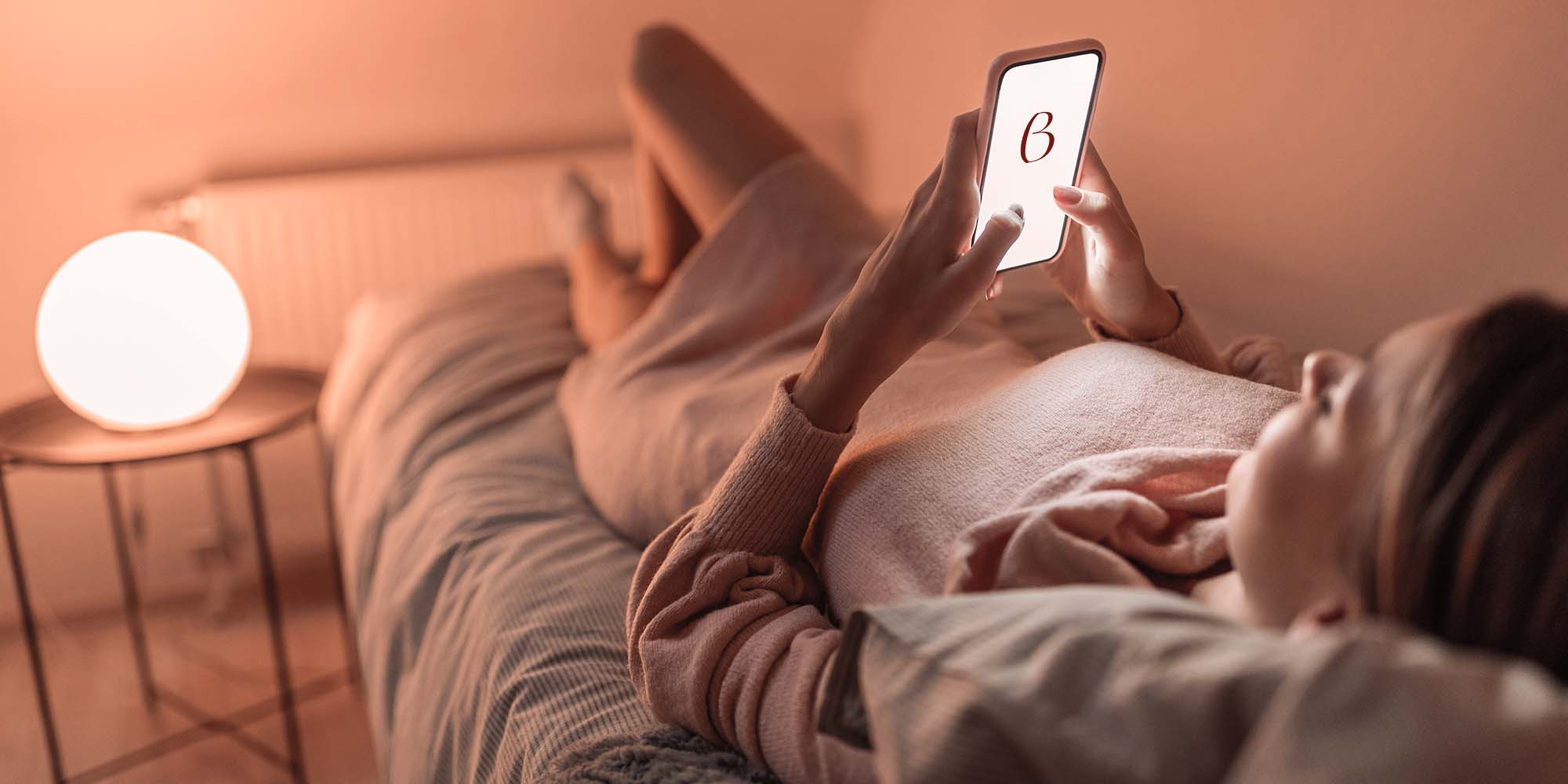 Una donna che utilizza il suo smartphone per iscriversi alla newsletter di Beliamo, con un letto elegante sullo sfondo.