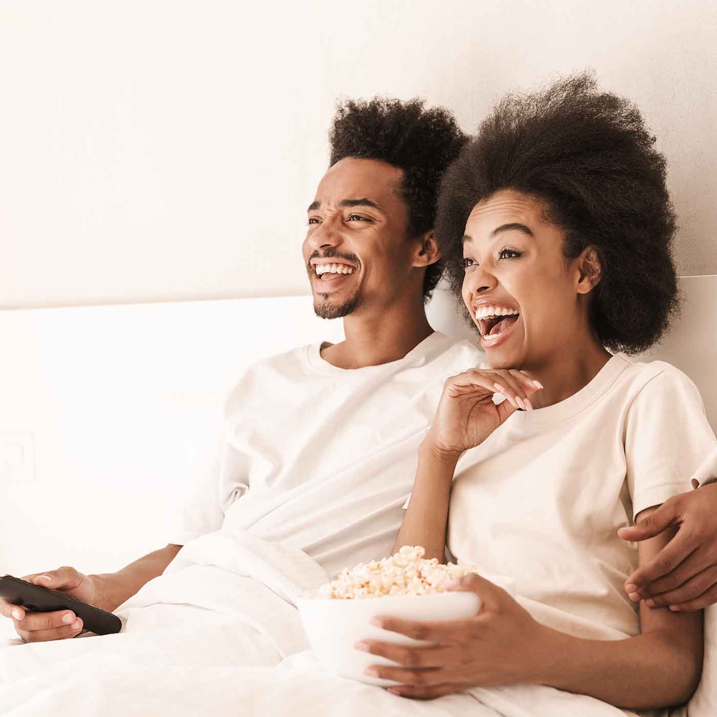 Ragazzi sorridenti nel proprio letto mentre mangiano pop corn guardando la tv by Beliamo